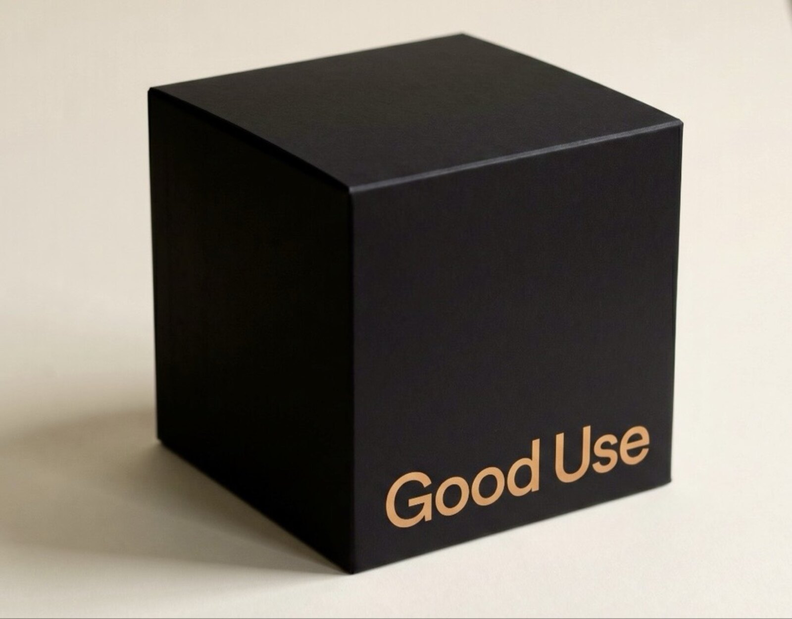A matte black cubic box with the Good Use wordmark in warm amber on the front face, on a cream backdrop in soft natural light.